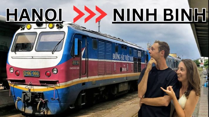 A couple standing in front of a blue and red Vietnamese train at a station, with text overlay "HANOI >> NINH BINH
